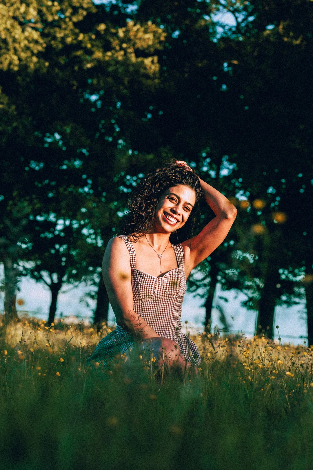 Smiling woman with curly hair in a gingham dress posing in a wildflower field under trees.