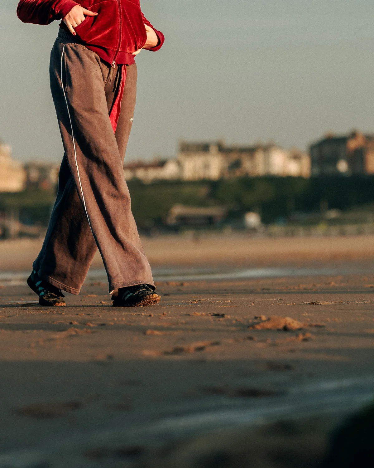 A person in a red velvet jacket and grey track pants walking on a sandy beach during golden hour.