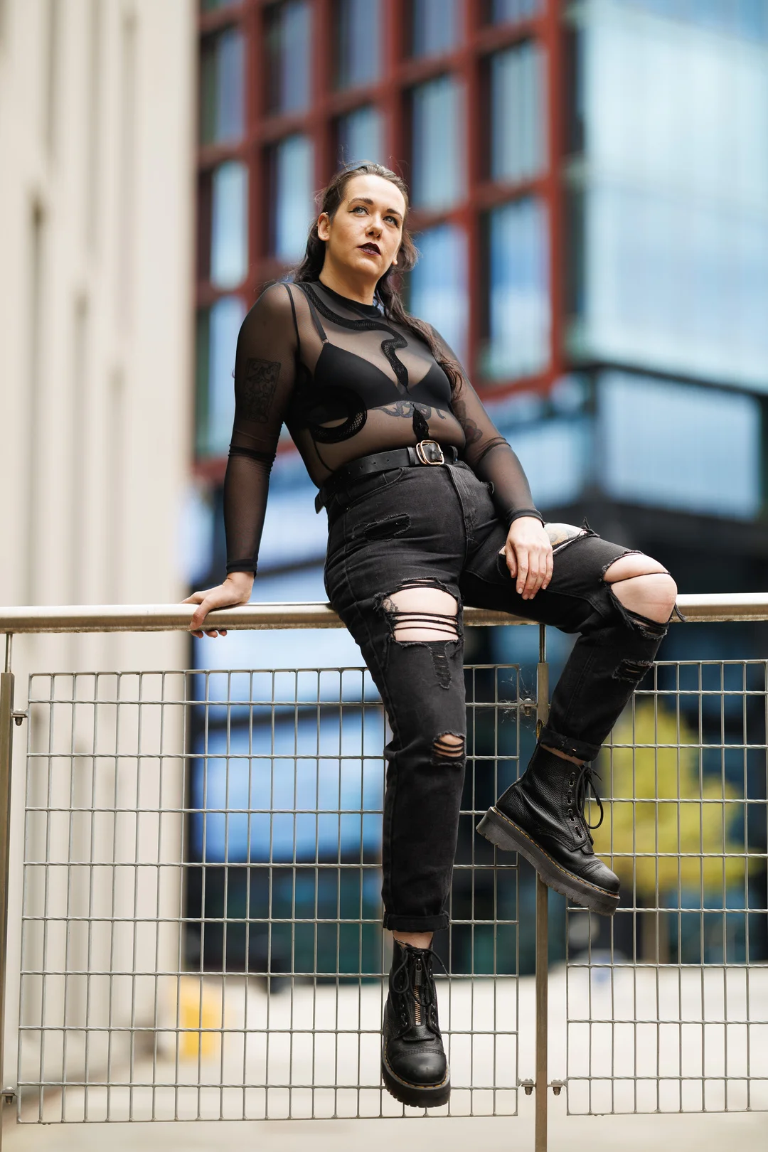 Full-body shot of a person in a sheer black top and ripped black jeans posing on an urban railing.