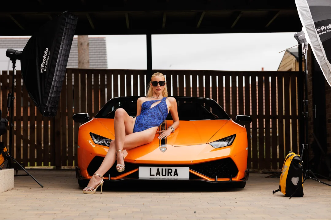 Woman in a blue swimsuit and sunglasses posing on the hood of an orange Lamborghini.