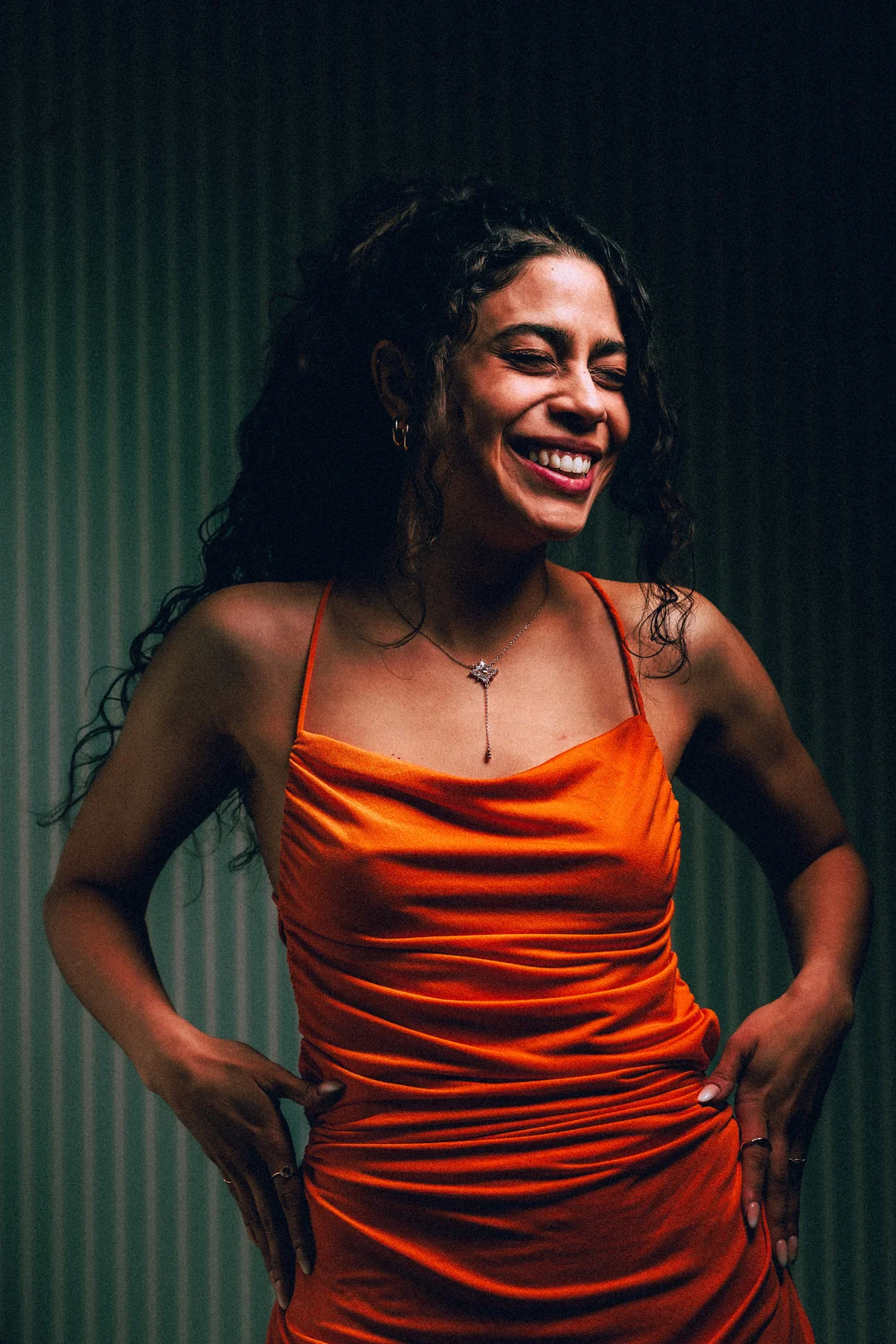 Smiling woman with curly hair in an orange satin dress against a dark textured background.