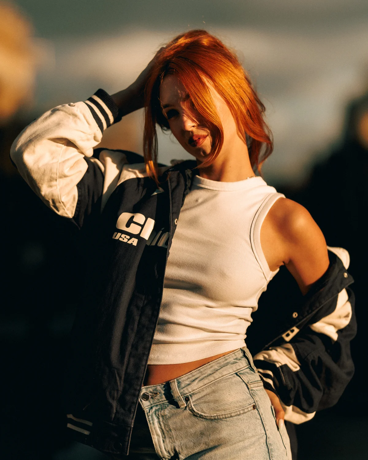 Young woman with red hair posing outdoors in a navy varsity jacket and white crop top during golden hour.