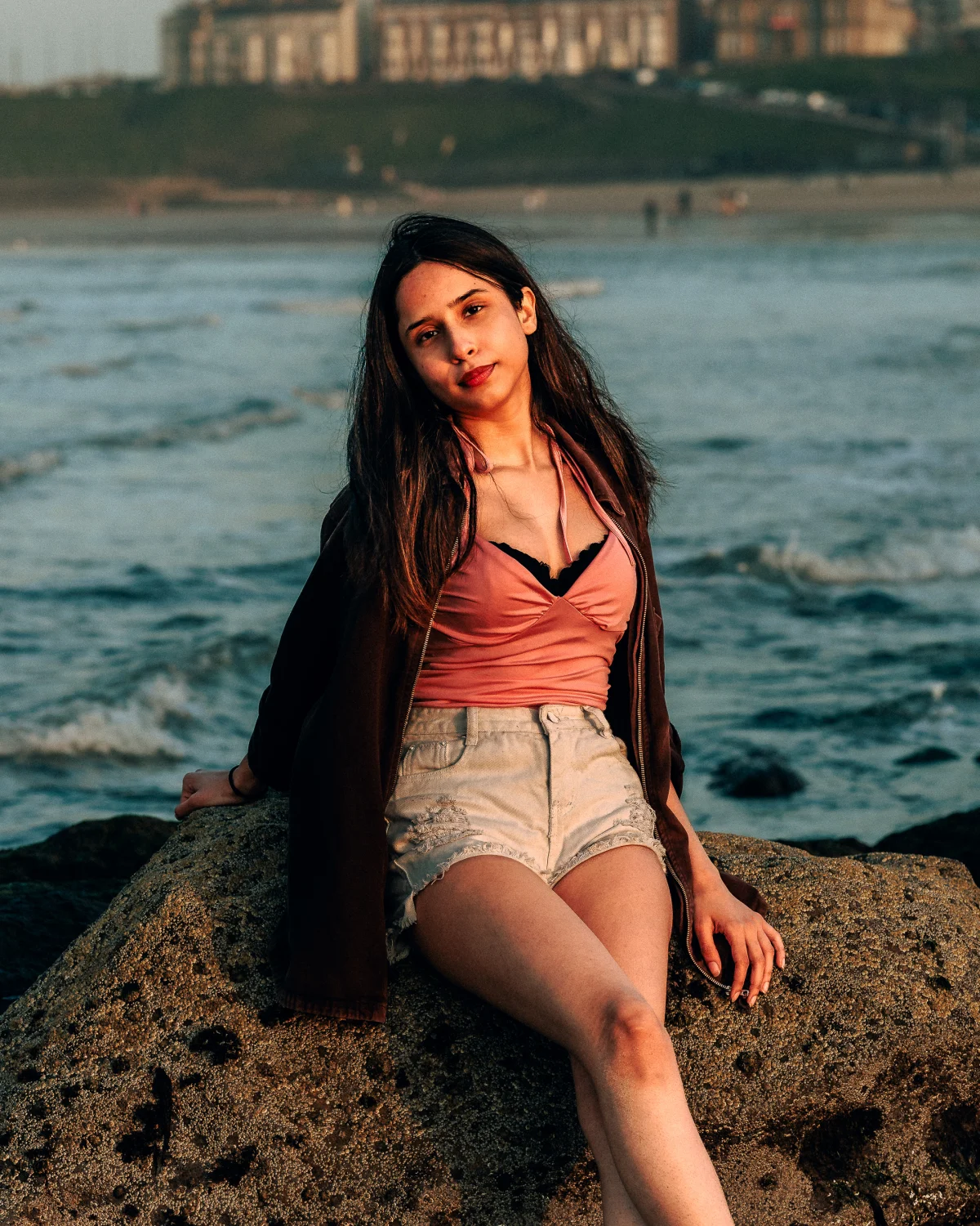 A young woman with long dark hair sitting on a rock by the ocean, wearing a pink top and white denim shorts.