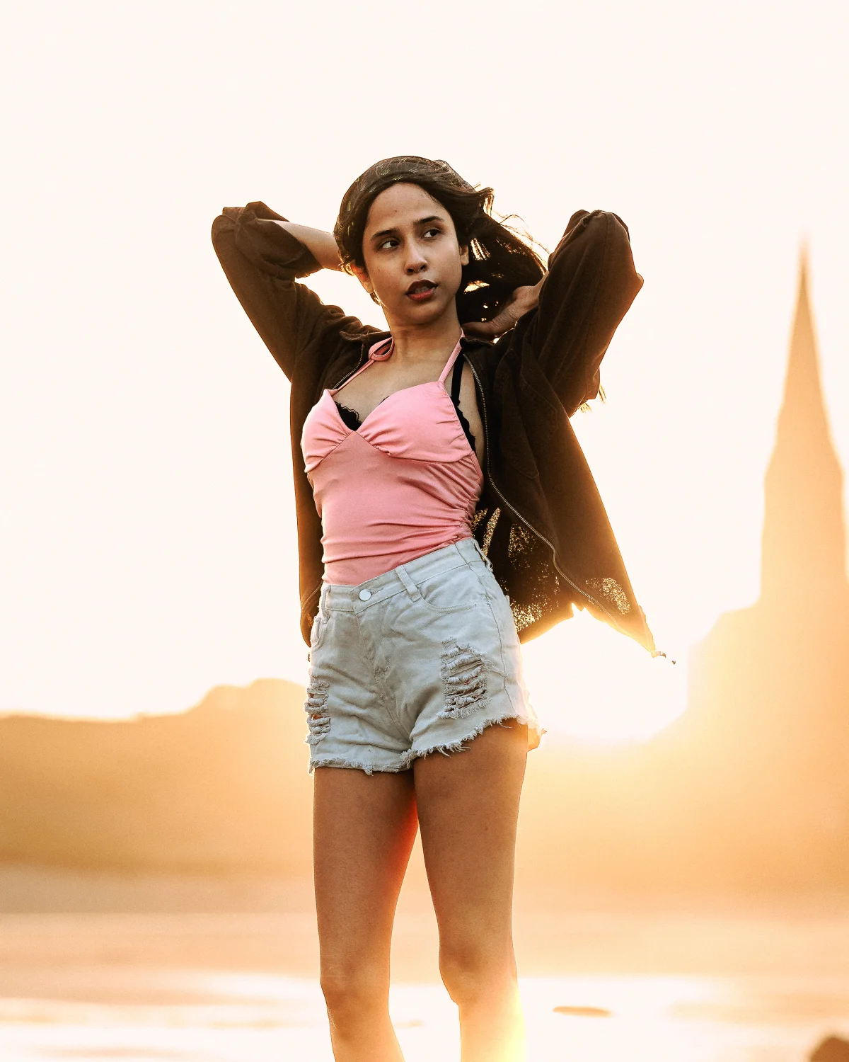 A woman posing in a pink top and denim shorts during a golden hour sunset.