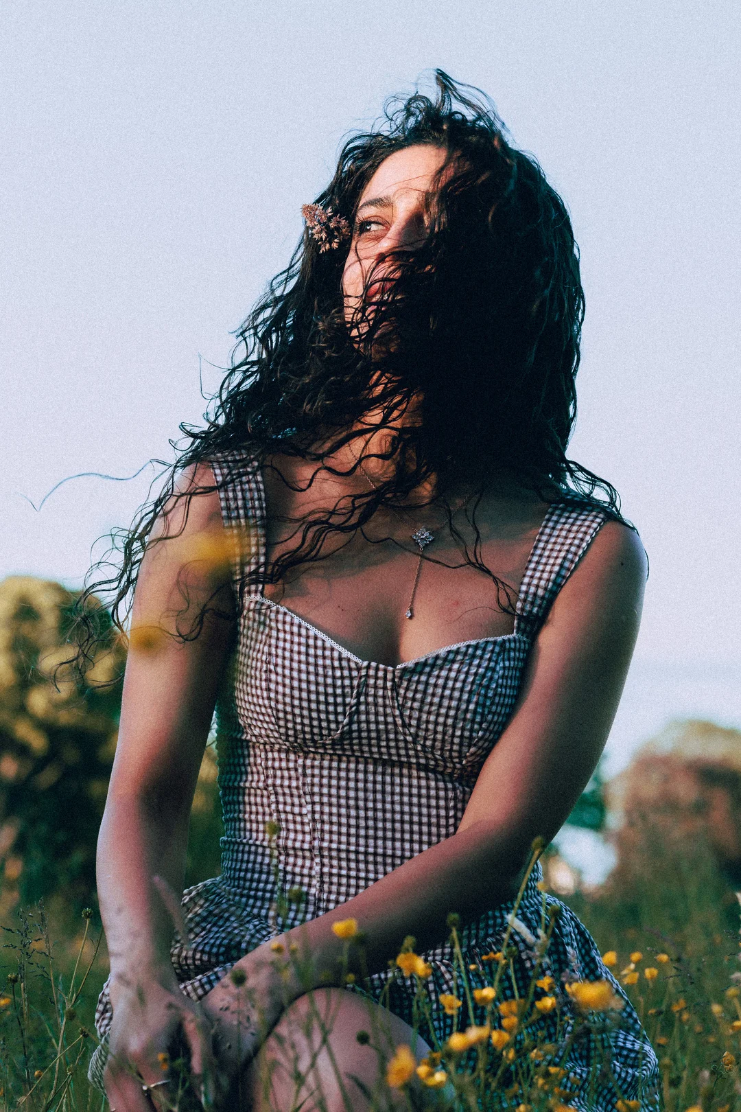 Woman with long curly hair in a gingham dress sitting in a field of yellow wildflowers.