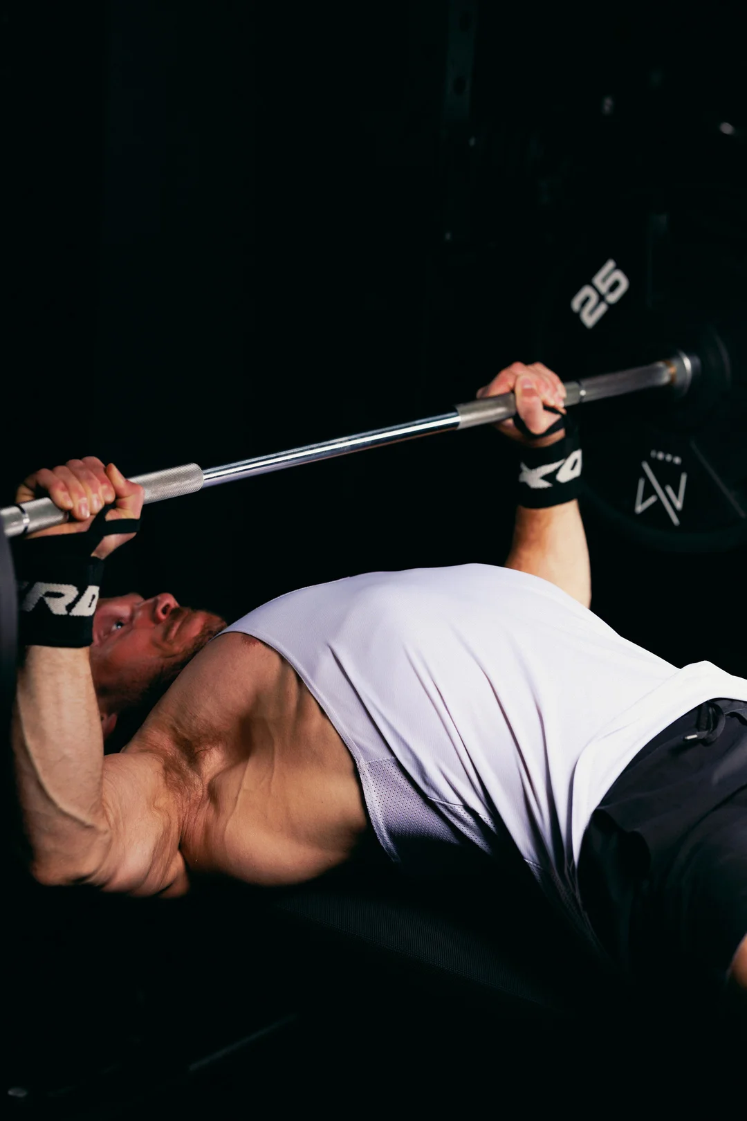 A muscular man performing a bench press with a barbell in a dark, moody gym setting.