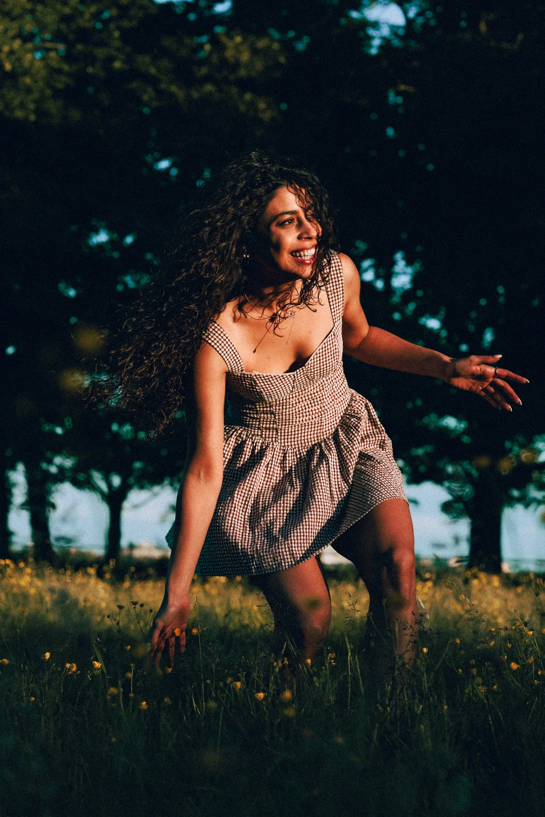 Smiling woman with curly hair in a gingham dress posing in a flower field at dusk.