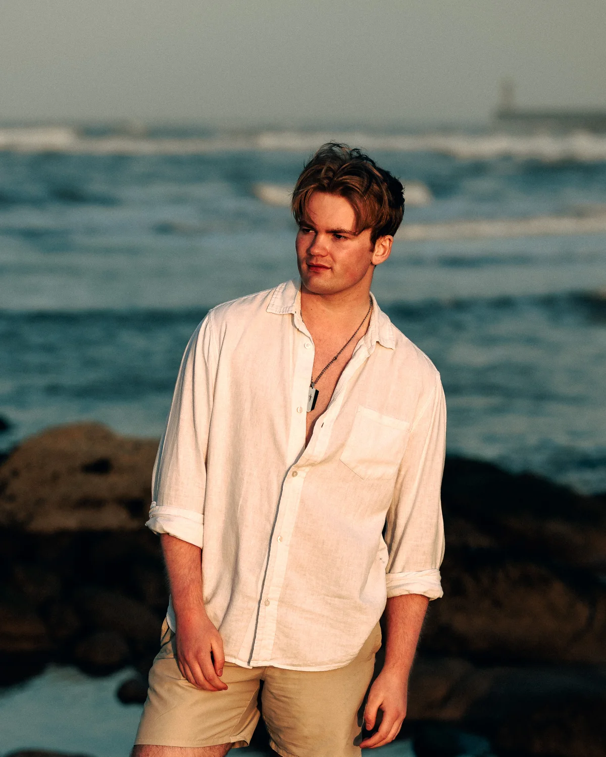 A young man in a white linen shirt and khaki shorts standing on a rocky beach during golden hour.