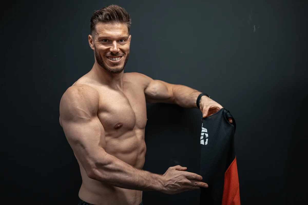 Muscular man smiling and holding a black and red athletic shirt against a dark studio background.