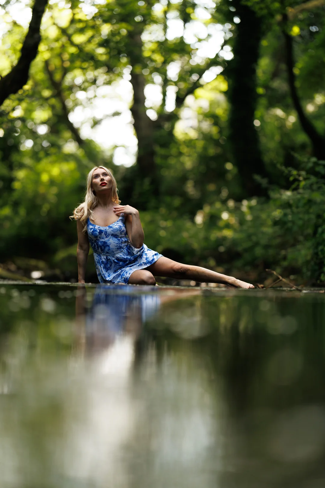 Blonde woman in a blue floral dress posing in a shallow forest stream with a reflection in the water.