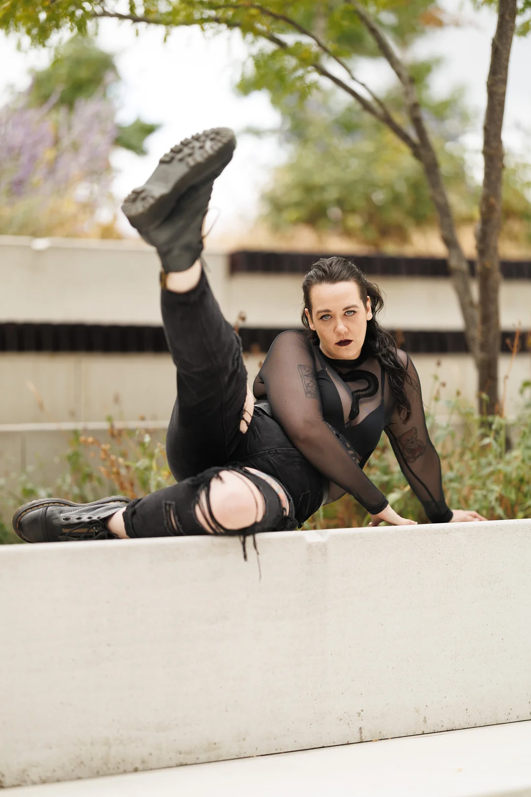 Person with dark hair and makeup posing on a concrete ledge outdoors in a black sheer top and ripped jeans.