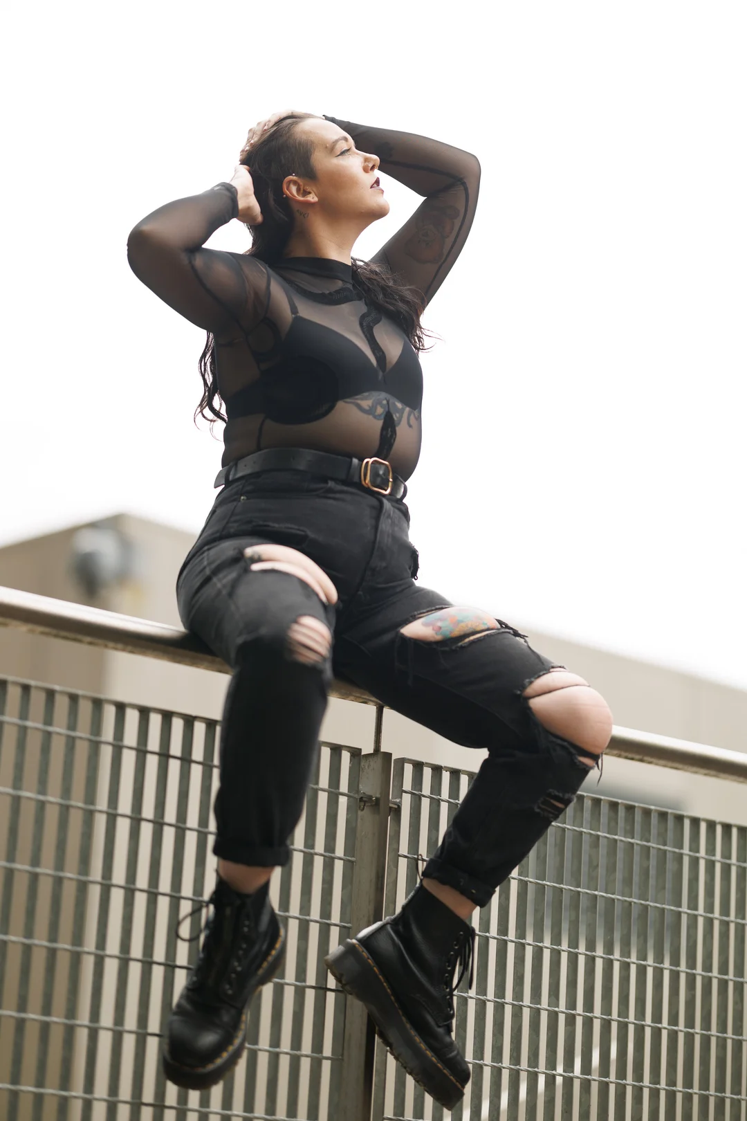 Woman in a black sheer top and ripped black jeans posing on a metal railing against a bright, overcast sky.