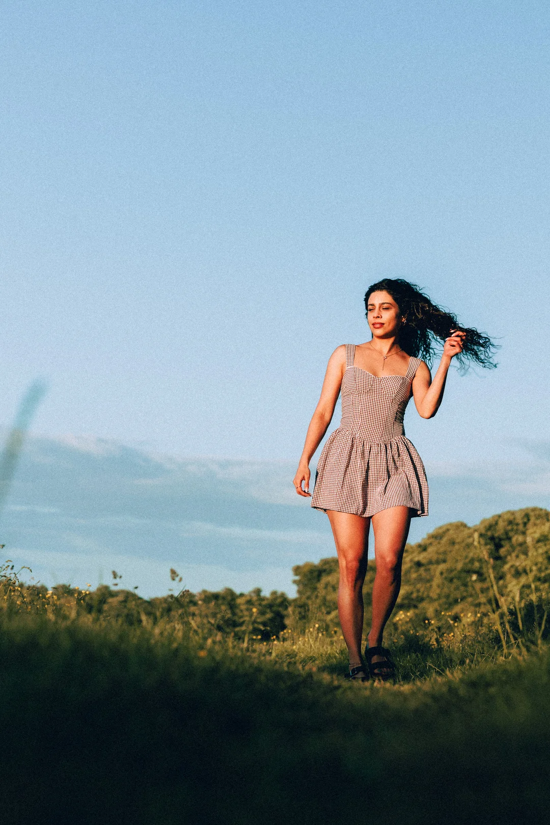 A woman with curly hair in a red and white gingham dress standing in a sunny grassy field.
