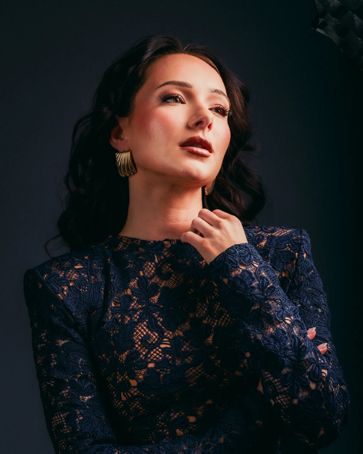 A woman with dark hair and red lipstick wearing a navy blue lace dress, posing against a dark background.