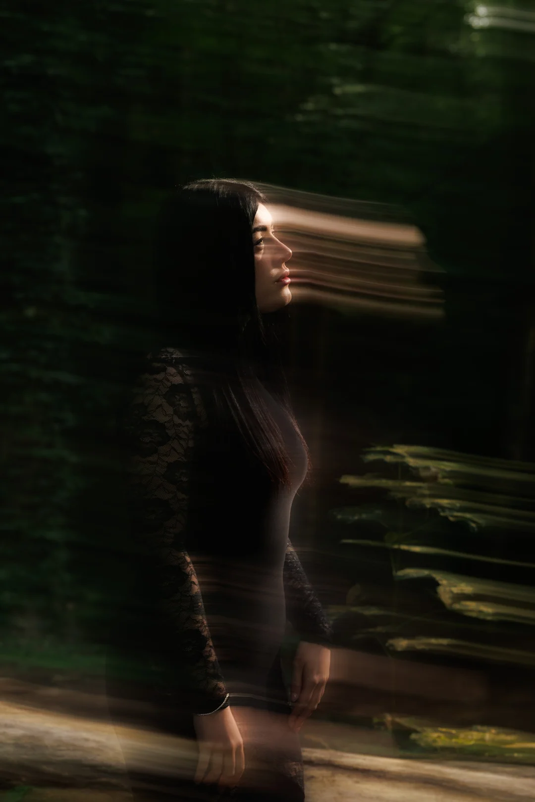 Moody creative portrait of a woman in a black lace outfit in a dark forest with motion blur.