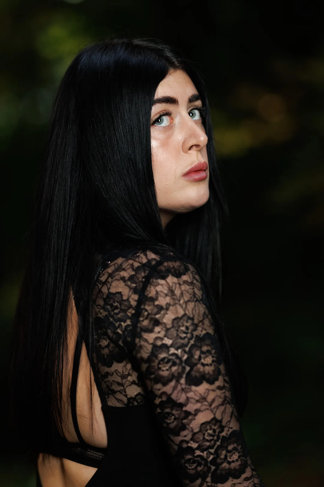 A woman with long black hair in a black lace dress looking over her shoulder against a dark blurred background.