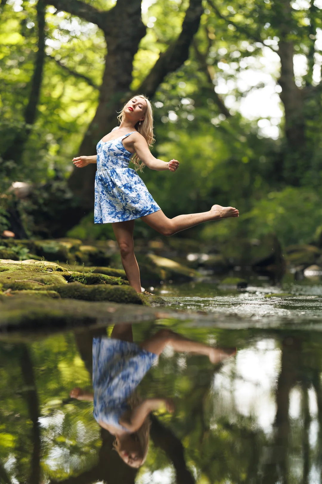 Woman in a blue floral dress posing on a mossy rock by a forest stream with a clear reflection in the water.