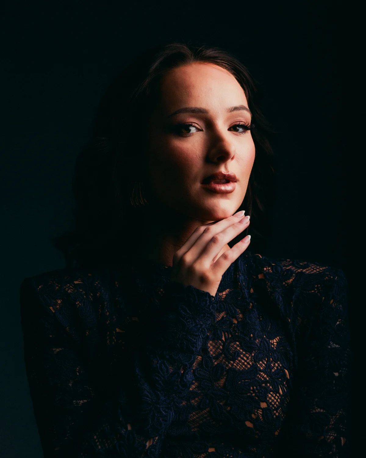 Moody portrait of a woman in a dark lace dress against a black background, hand touching her chin.