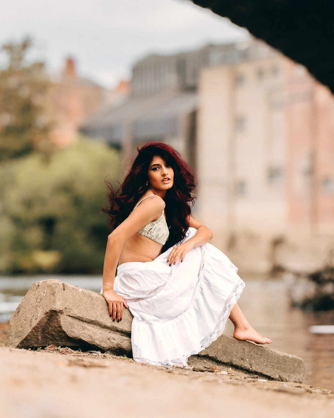 Woman with red hair in a white skirt and bralette sitting on a rock by a river with a city backdrop.