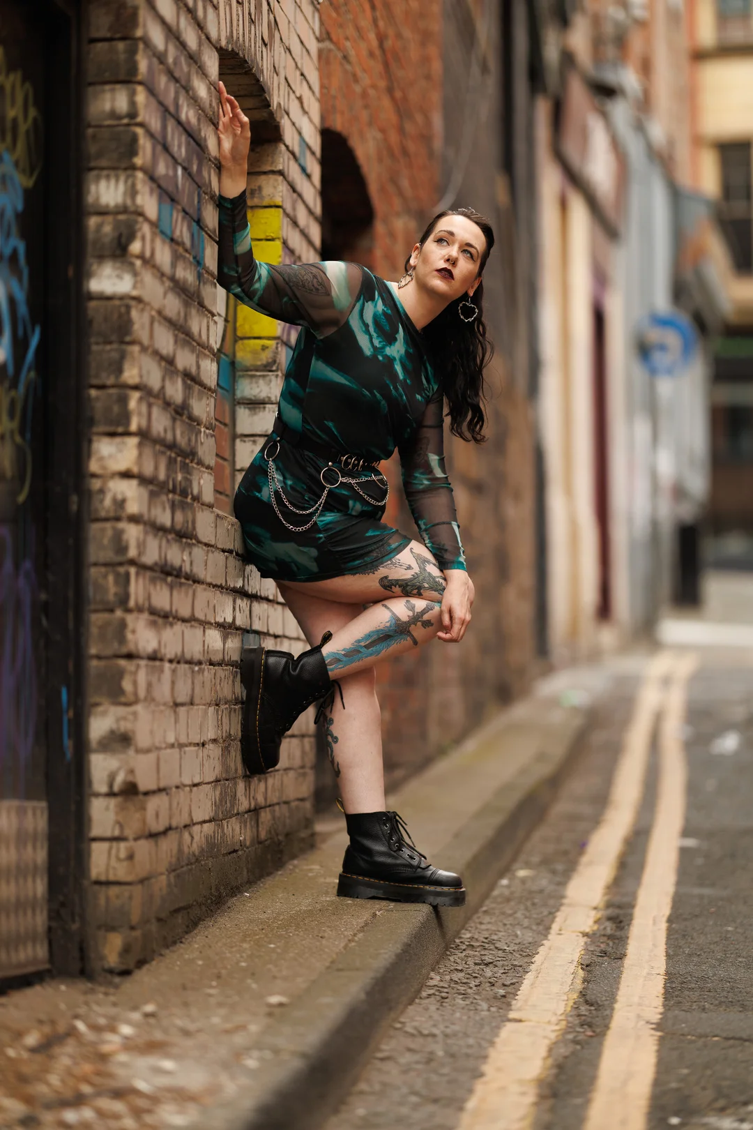Woman with tattoos in a green patterned dress and black boots posing against a brick wall in an urban alley.
