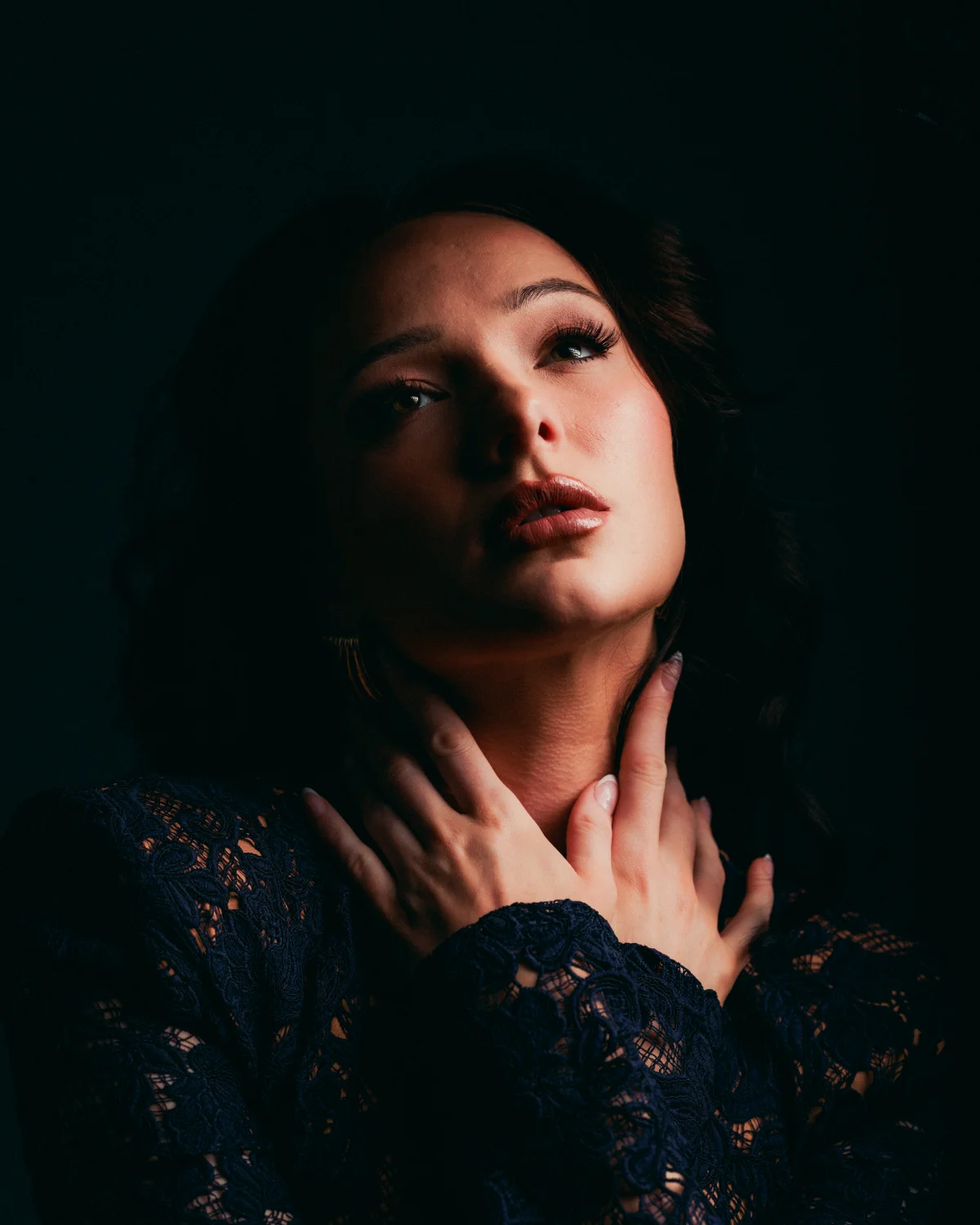Moody close-up portrait of a woman in a dark lace dress with her hands on her neck against a black background.