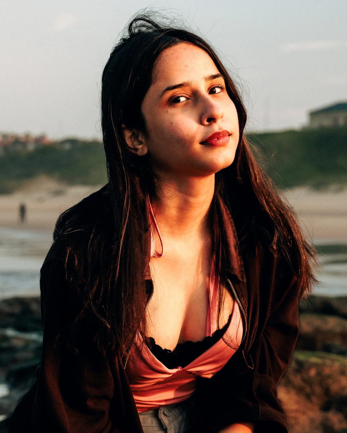 Close-up portrait of a woman with long dark hair at a beach during golden hour, wearing a pink top and dark jacket.