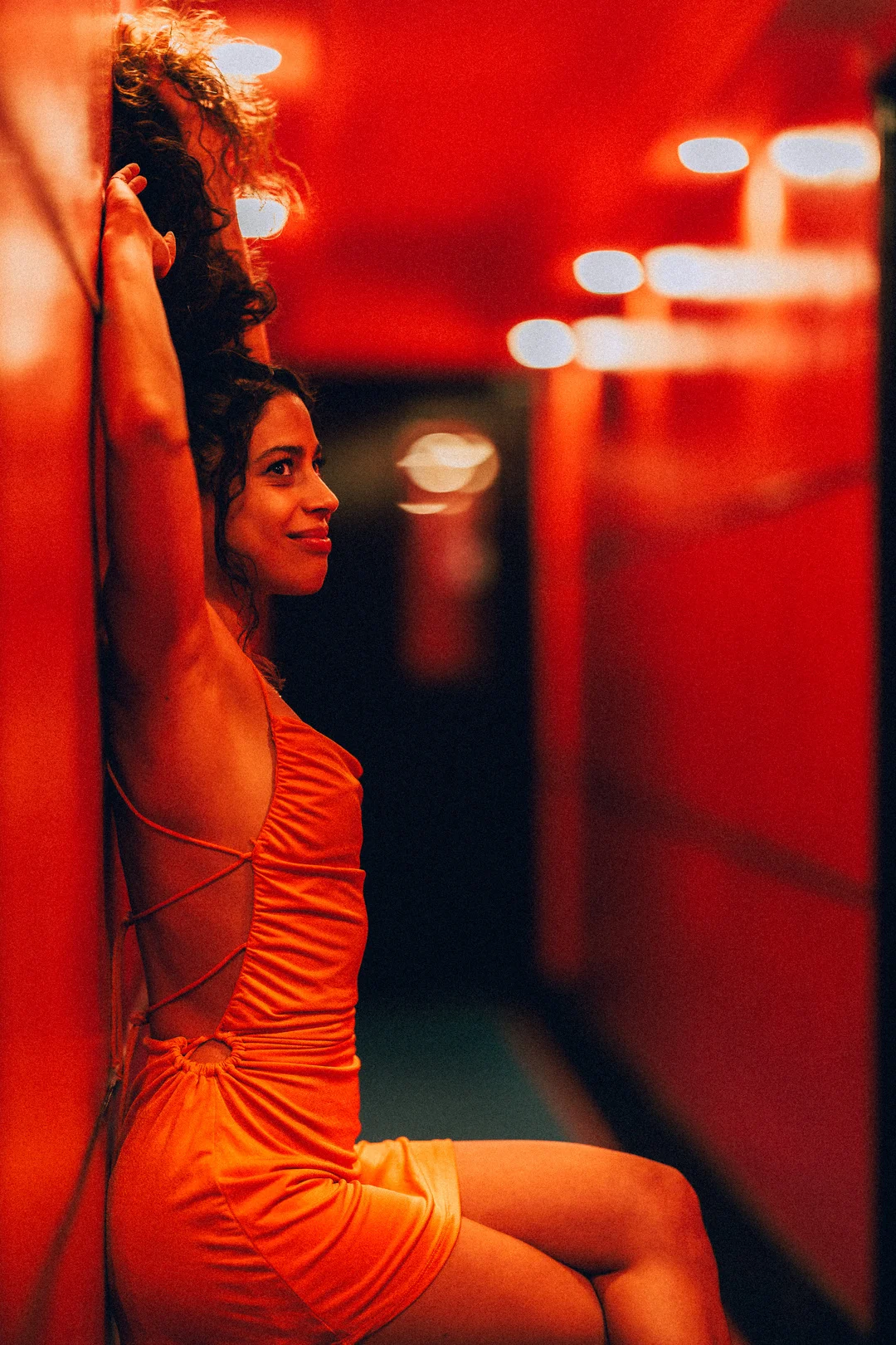 Woman in a bright orange backless dress posing in a moody, red-lit corridor.
