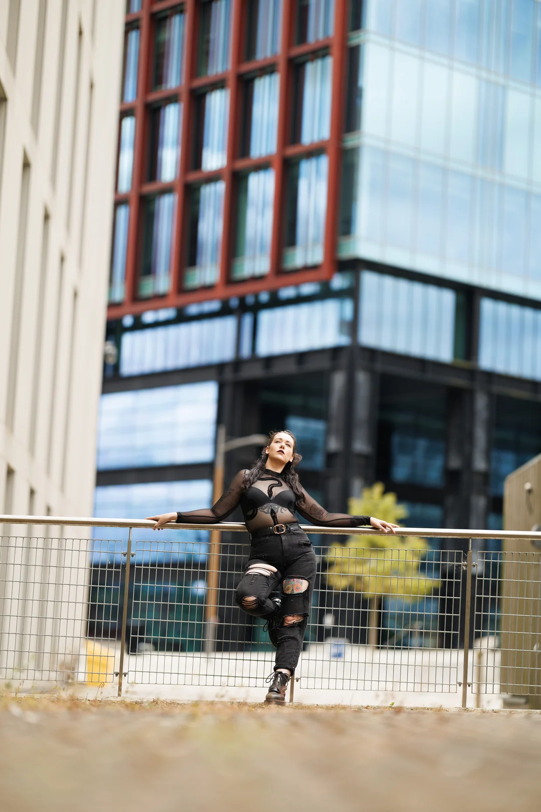 Woman in black mesh top and ripped jeans posing by a railing in an urban city setting.