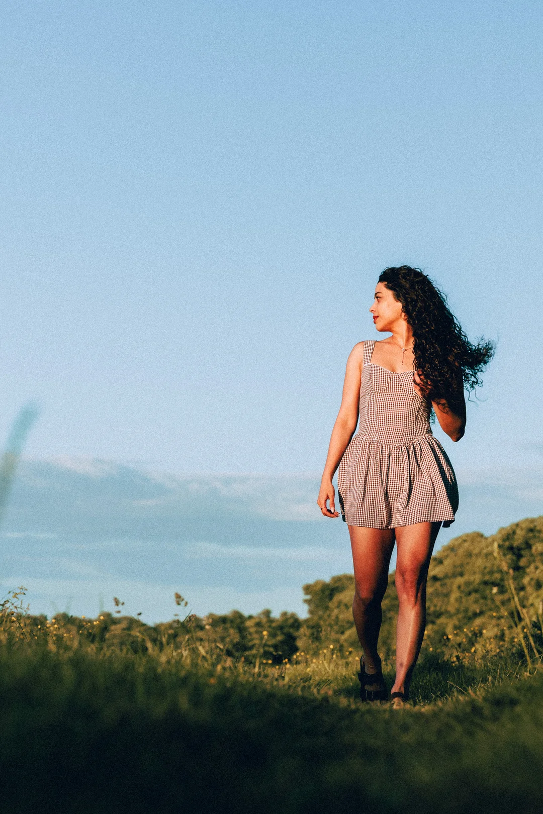 A woman with curly hair in a red and white gingham dress standing in a grassy field under a clear blue sky.