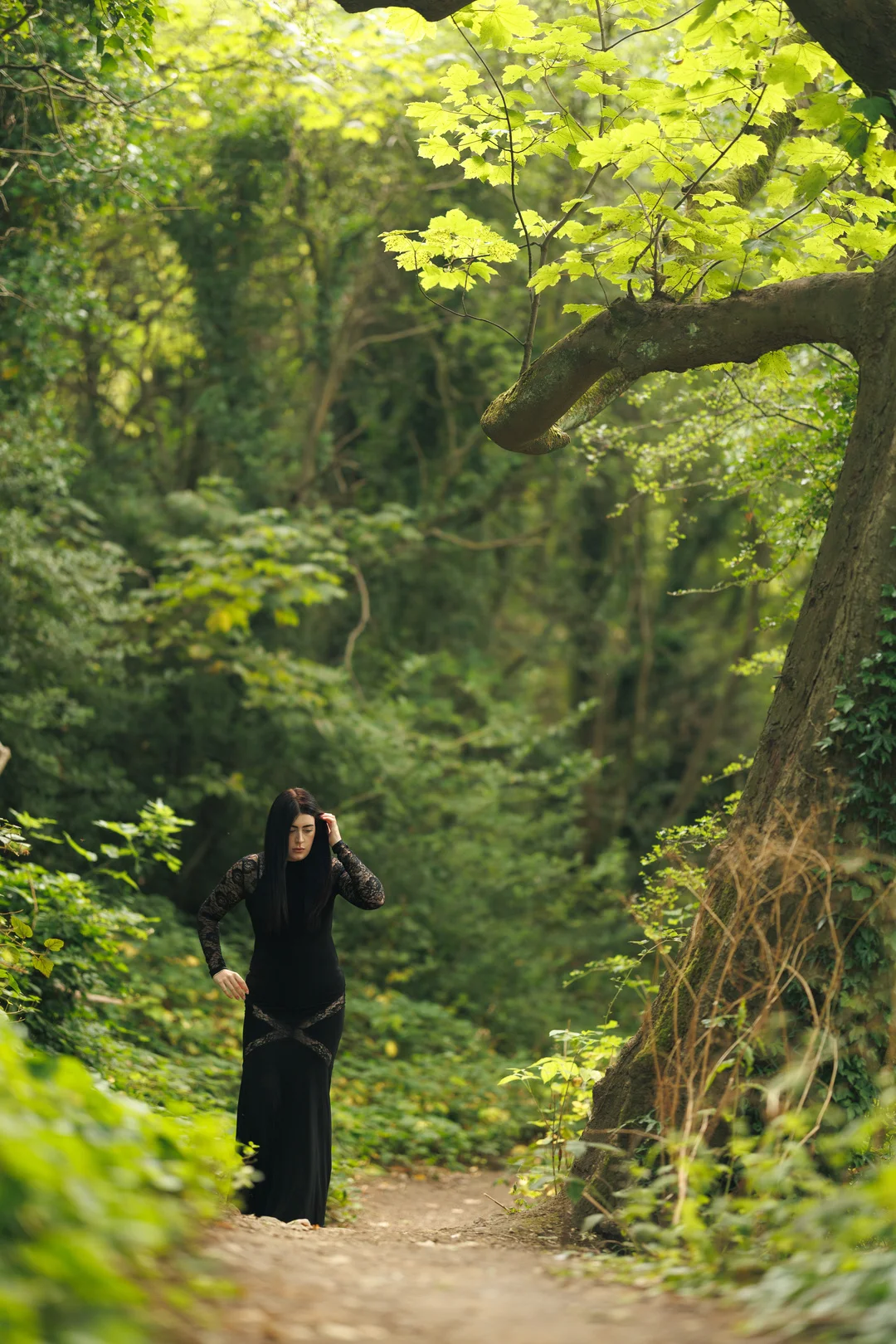 A woman in a long black lace dress standing on a path in a lush green forest.