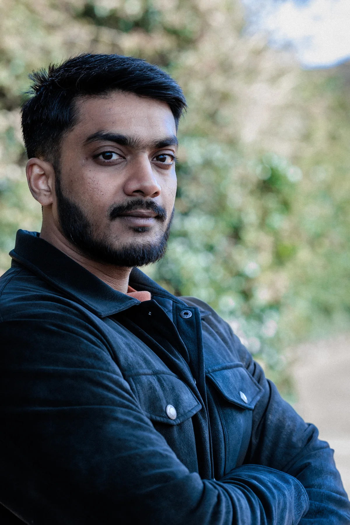 Close-up portrait of a man with a beard wearing a black jacket against a blurred outdoor background.