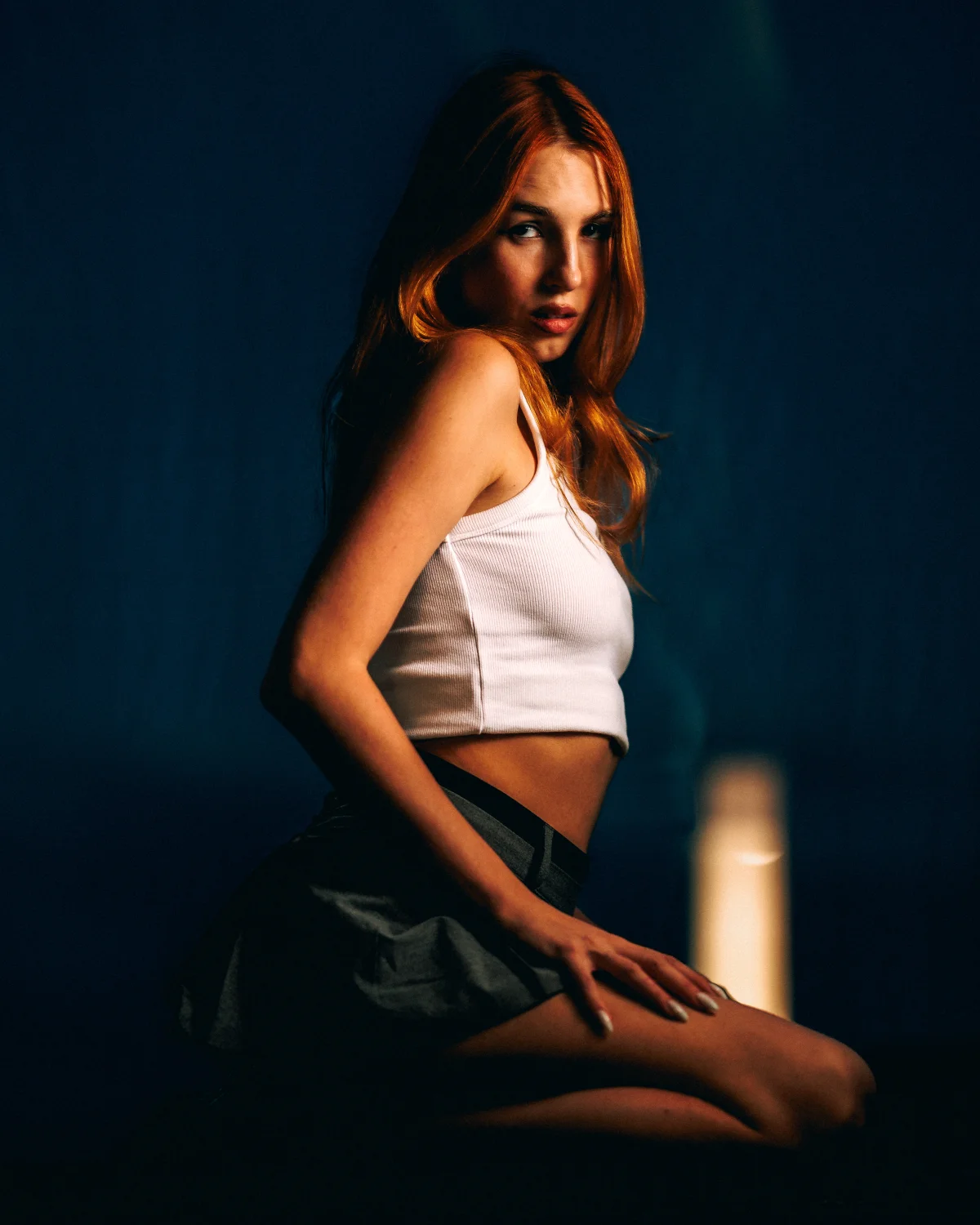 Moody studio portrait of a red-haired woman in a white crop top and dark skirt against a deep blue background.