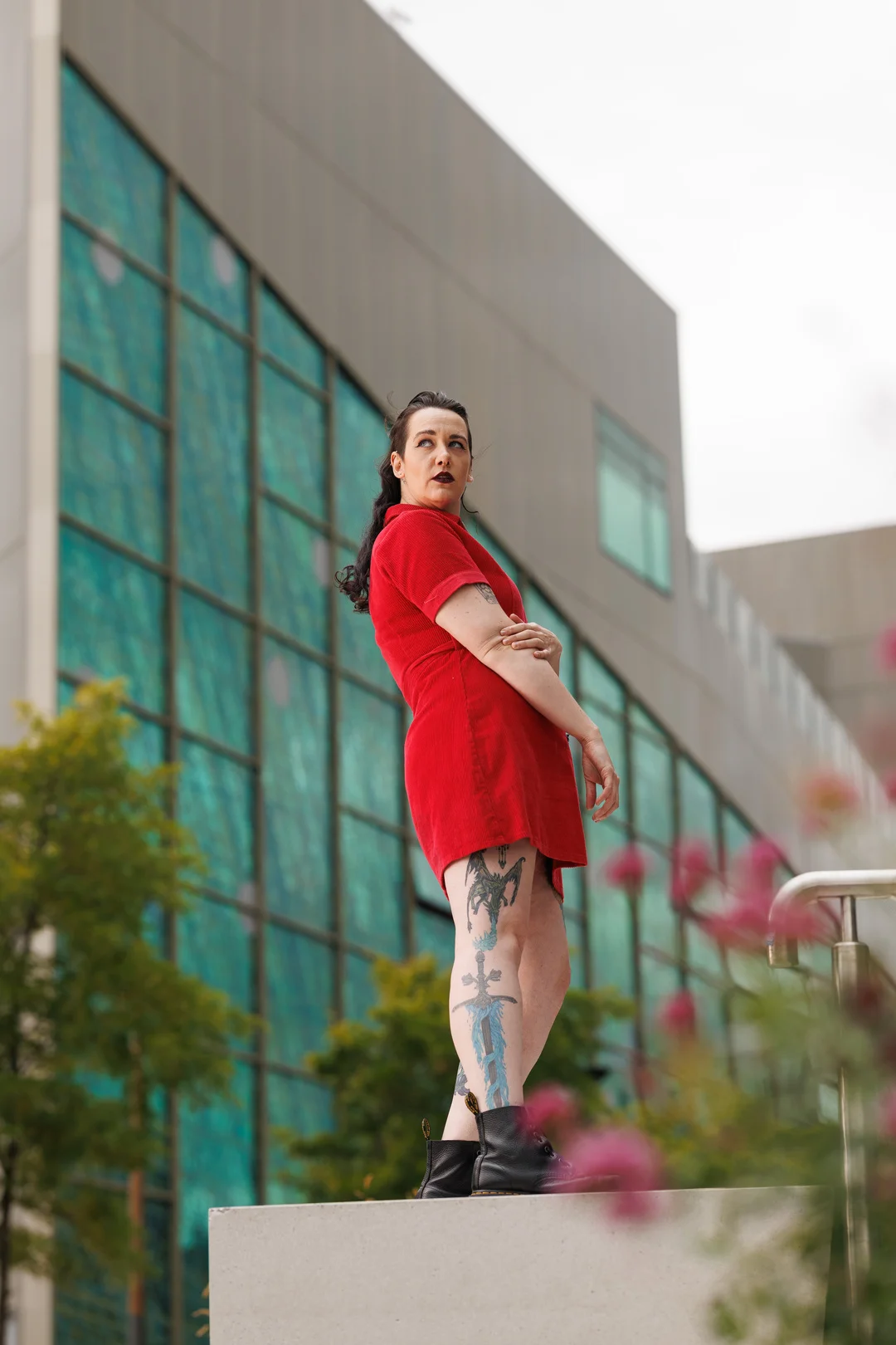 A woman in a short red dress with leg tattoos posing in front of a modern glass building.