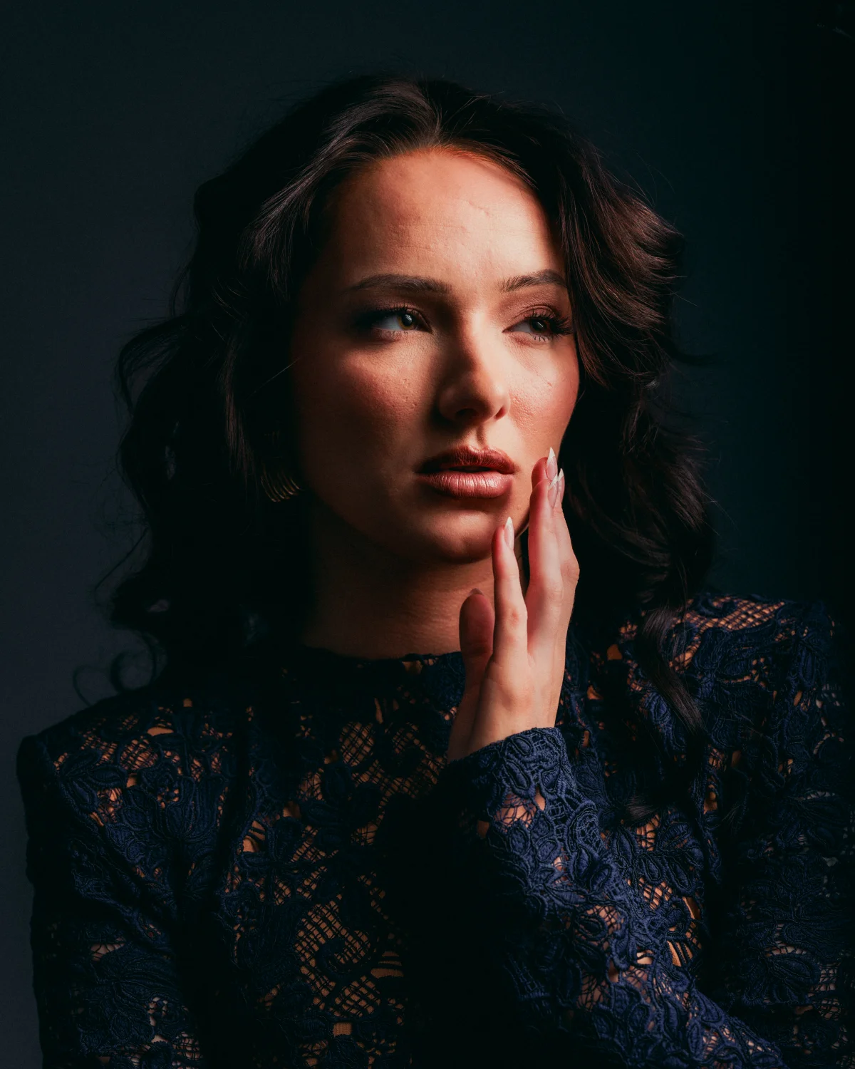 Moody close-up portrait of a woman with dark wavy hair and a navy lace dress against a dark background.