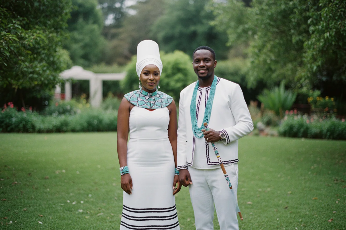 A Xhosa bride and groom in matching white umbhaco with black braid trim and turquoise Xhosa beadwork stand in an Eastern Cape garden