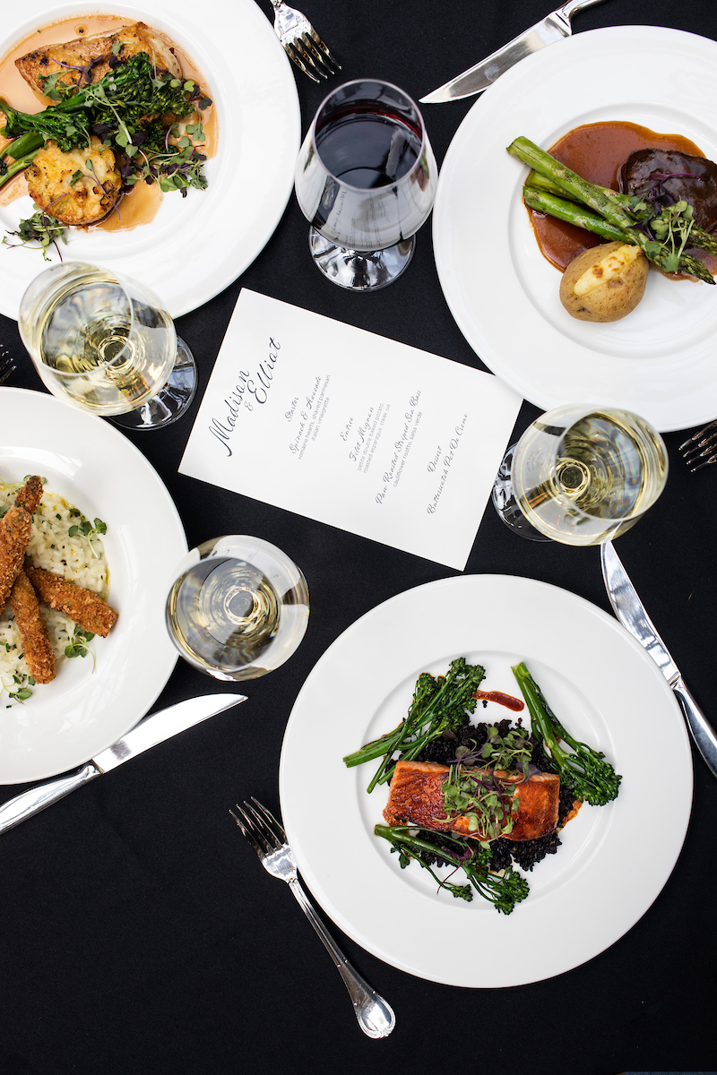 Four dishes of food on a black table cloth includes: red and white wine, salmon, chicken, filet and asparagus  