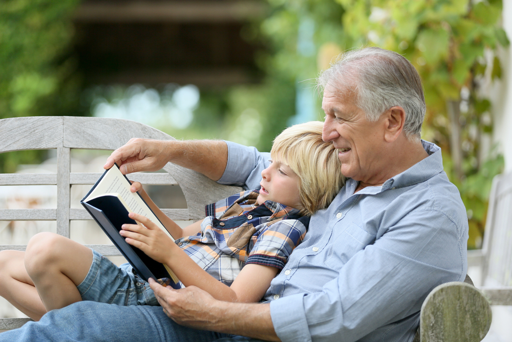 A grandson sits on his grandfather's lap while the grandpa reads a story to him