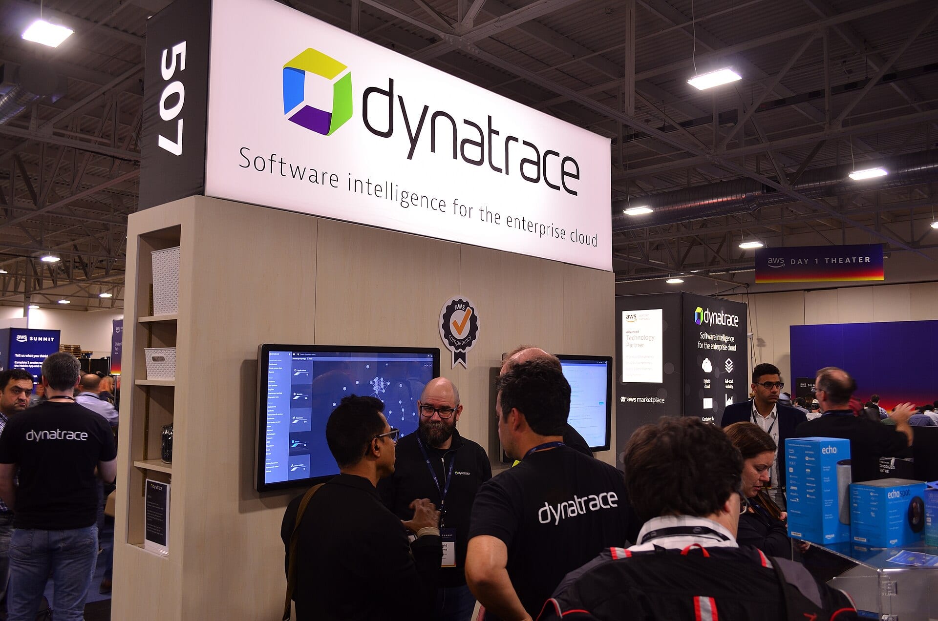 Dynatrace staff at the company's booth on the AWS Summit expo floor in Toronto, with screens running the Dynatrace dashboard