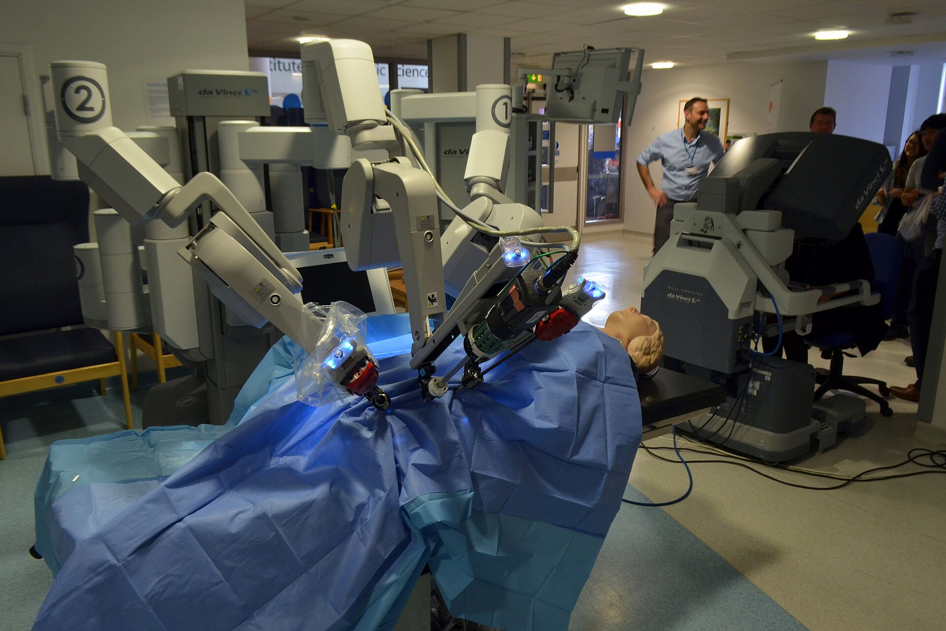 A da Vinci surgical system with its instrument arms over an operating table and a surgeon console at right