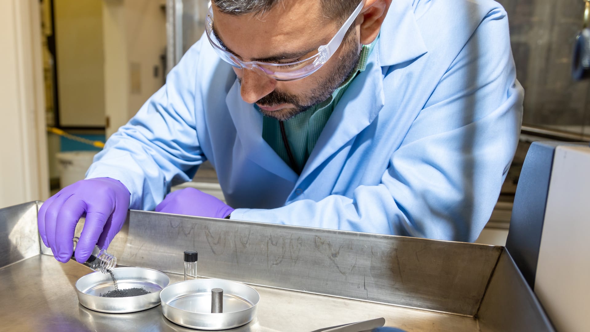A researcher in safety glasses and purple gloves pours TRISO nuclear fuel particles into a steel tray at Oak Ridge National Laboratory