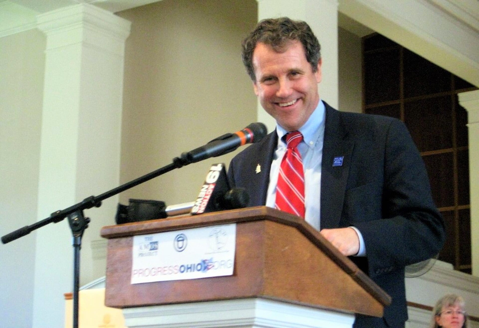 Sherrod Brown, smiling, speaks at a wooden podium with microphones at an event in Cleveland Heights, Ohio