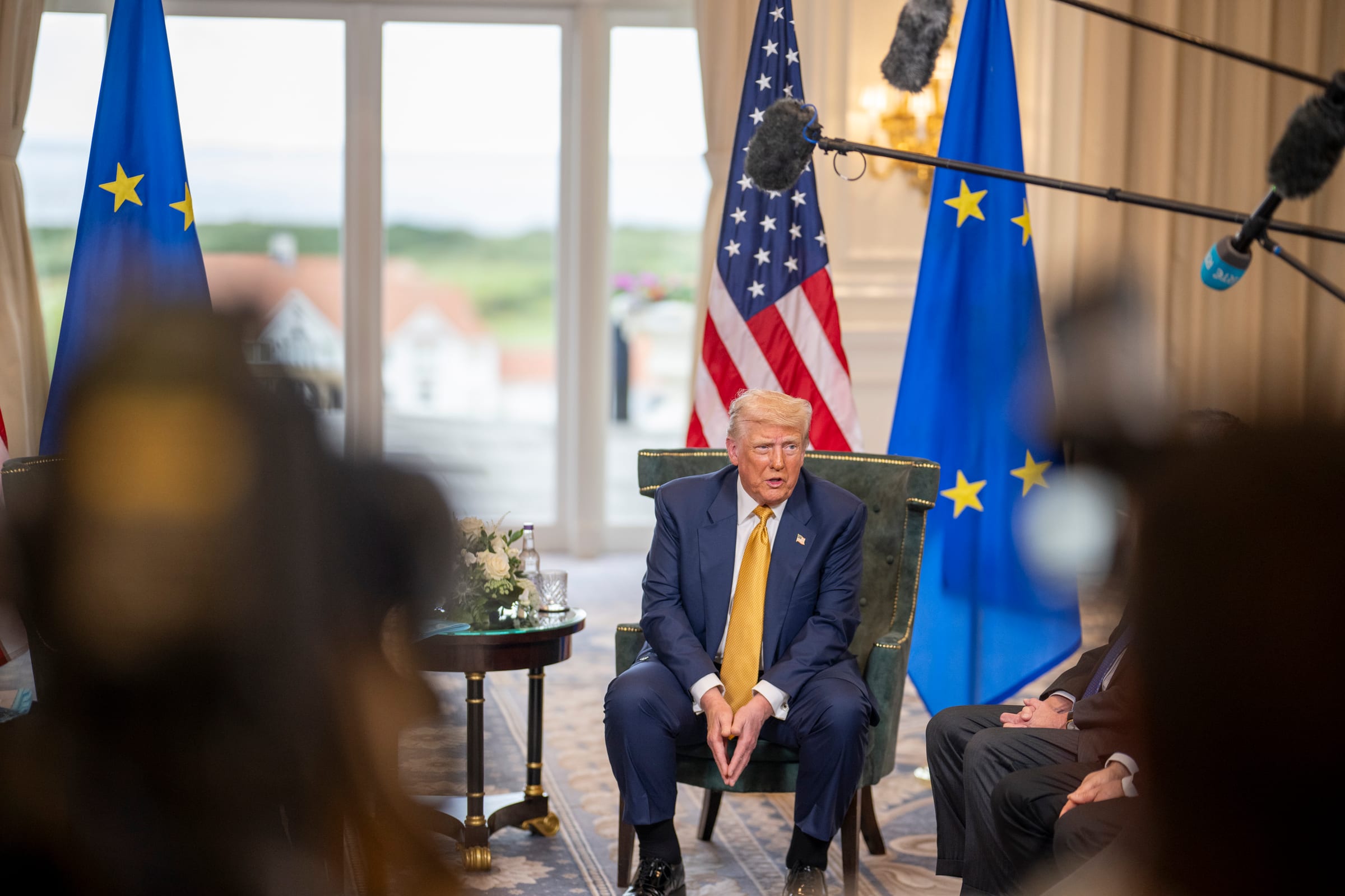 President Trump seated and speaking to reporters, with American and European Union flags behind him and microphone booms overhead