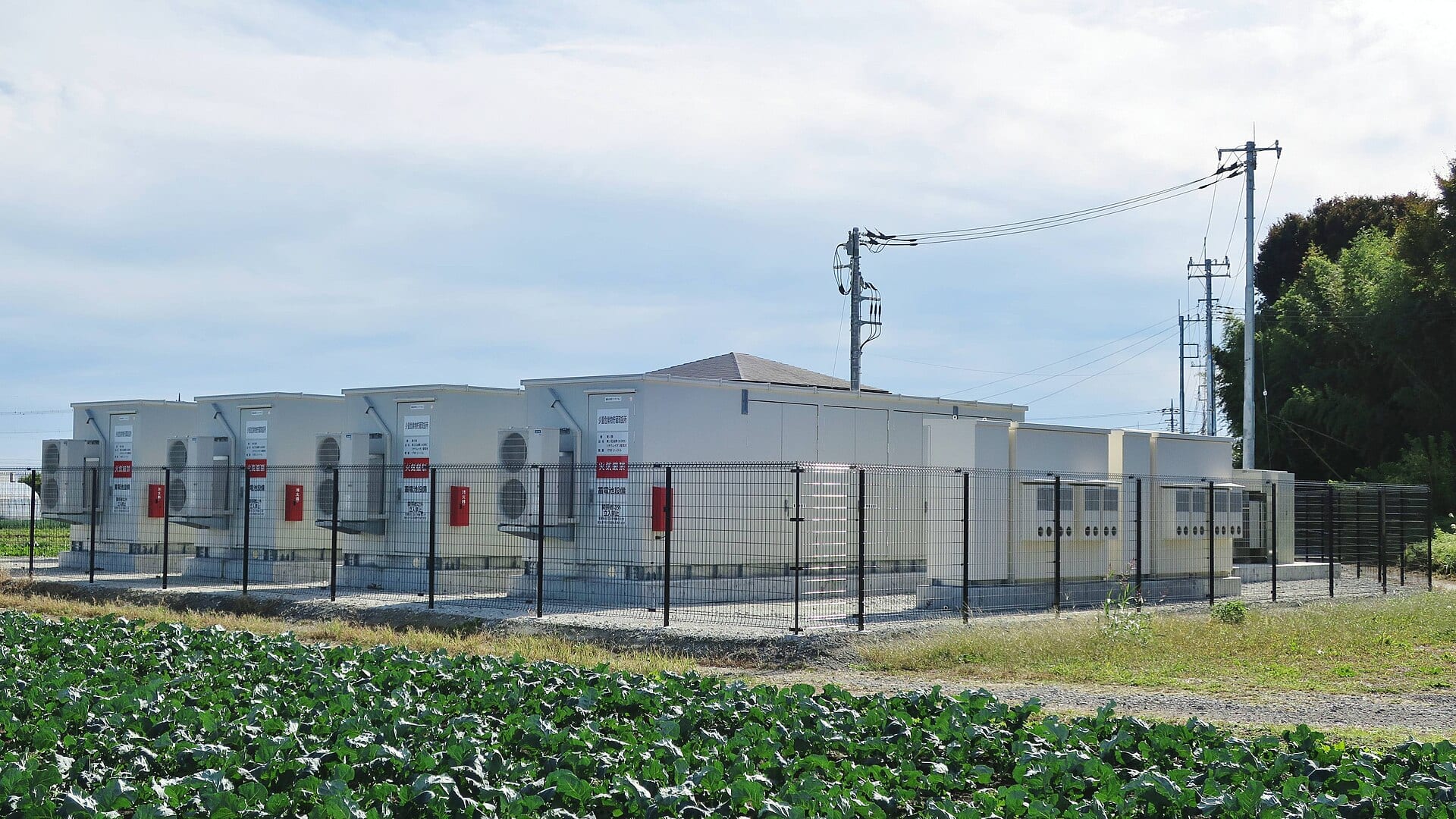 Containerized battery storage units behind fencing at a grid-scale battery storage power station in Japan, with power lines overhead.