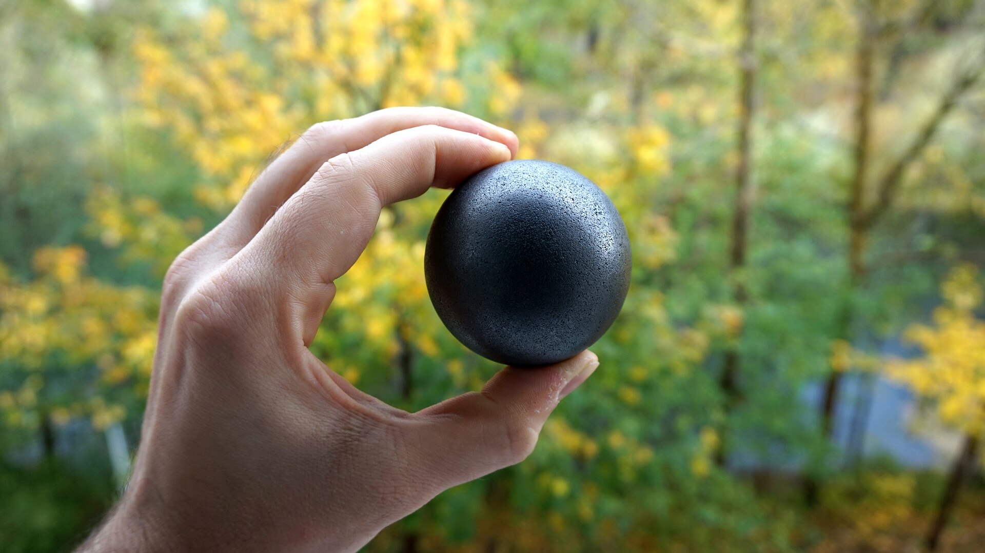 A hand holds a black graphite fuel pebble for a pebble-bed nuclear reactor, 60 millimetres across