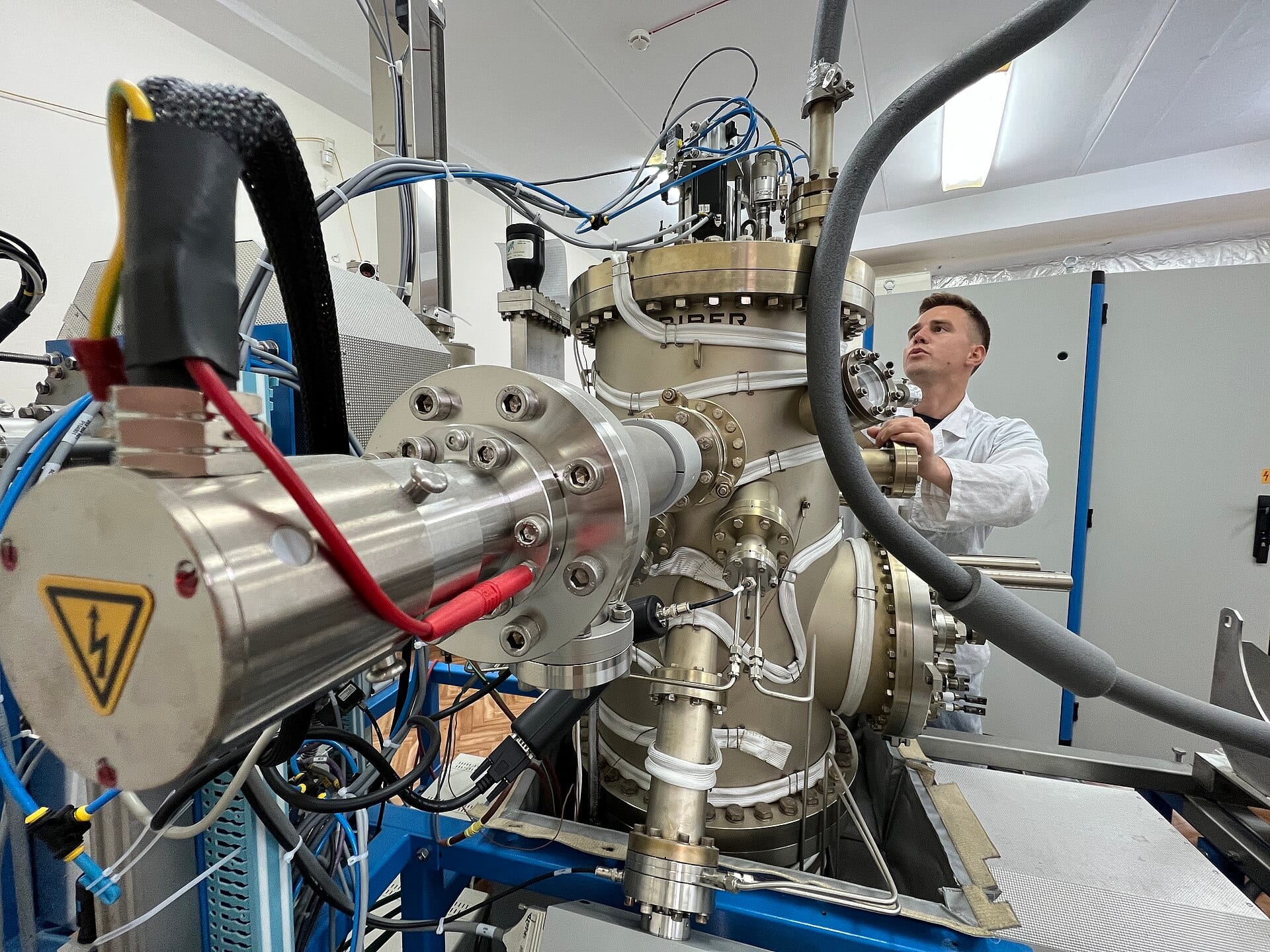 A technician in a white lab coat works on a Riber SIVA 21 molecular beam epitaxy system, a large steel vacuum chamber ringed with pipes, cables and flanges.