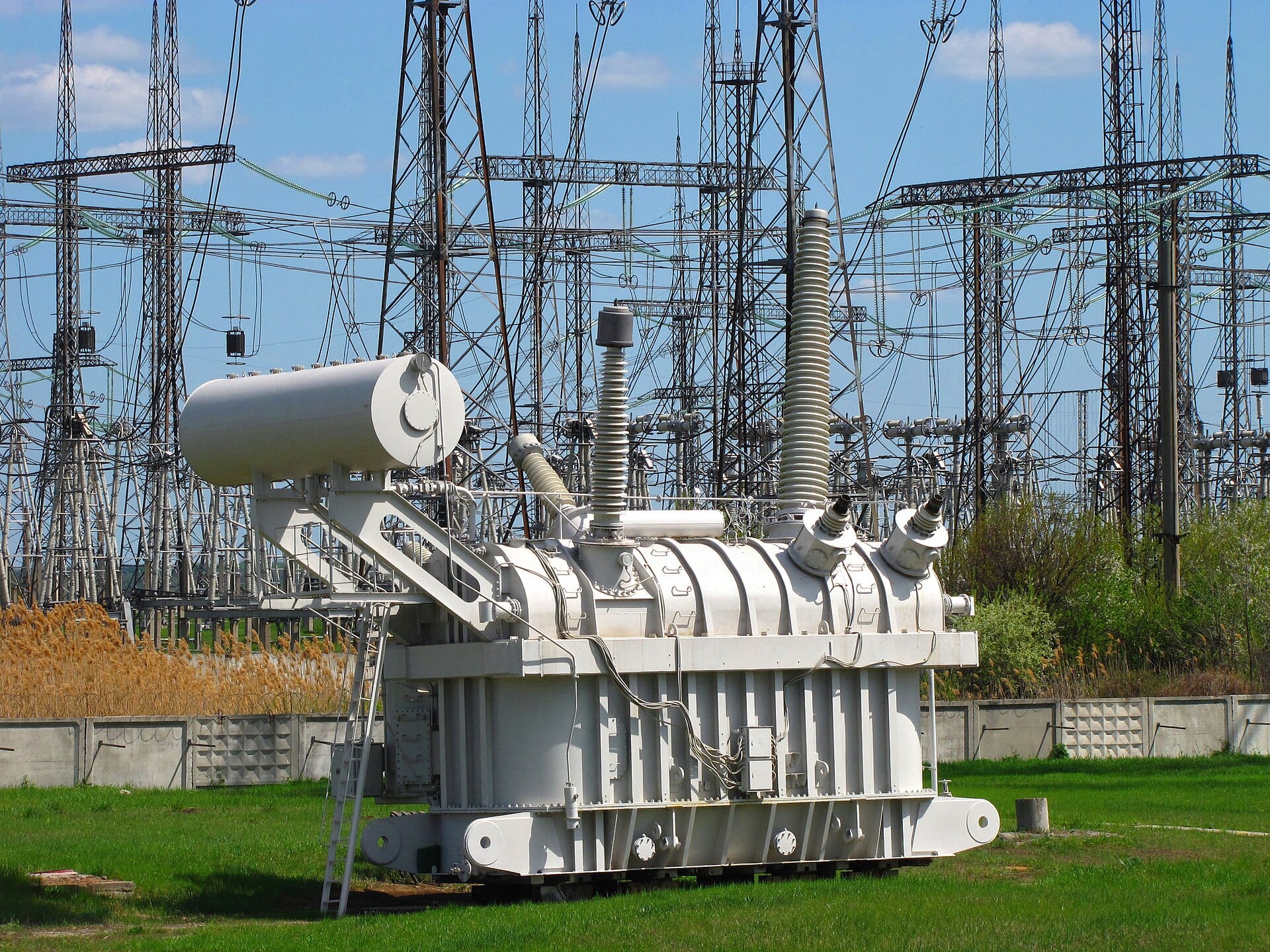 A 750 kV high-voltage transformer with tall ribbed bushings on top, standing in a substation surrounded by transmission gantries and overhead lines.