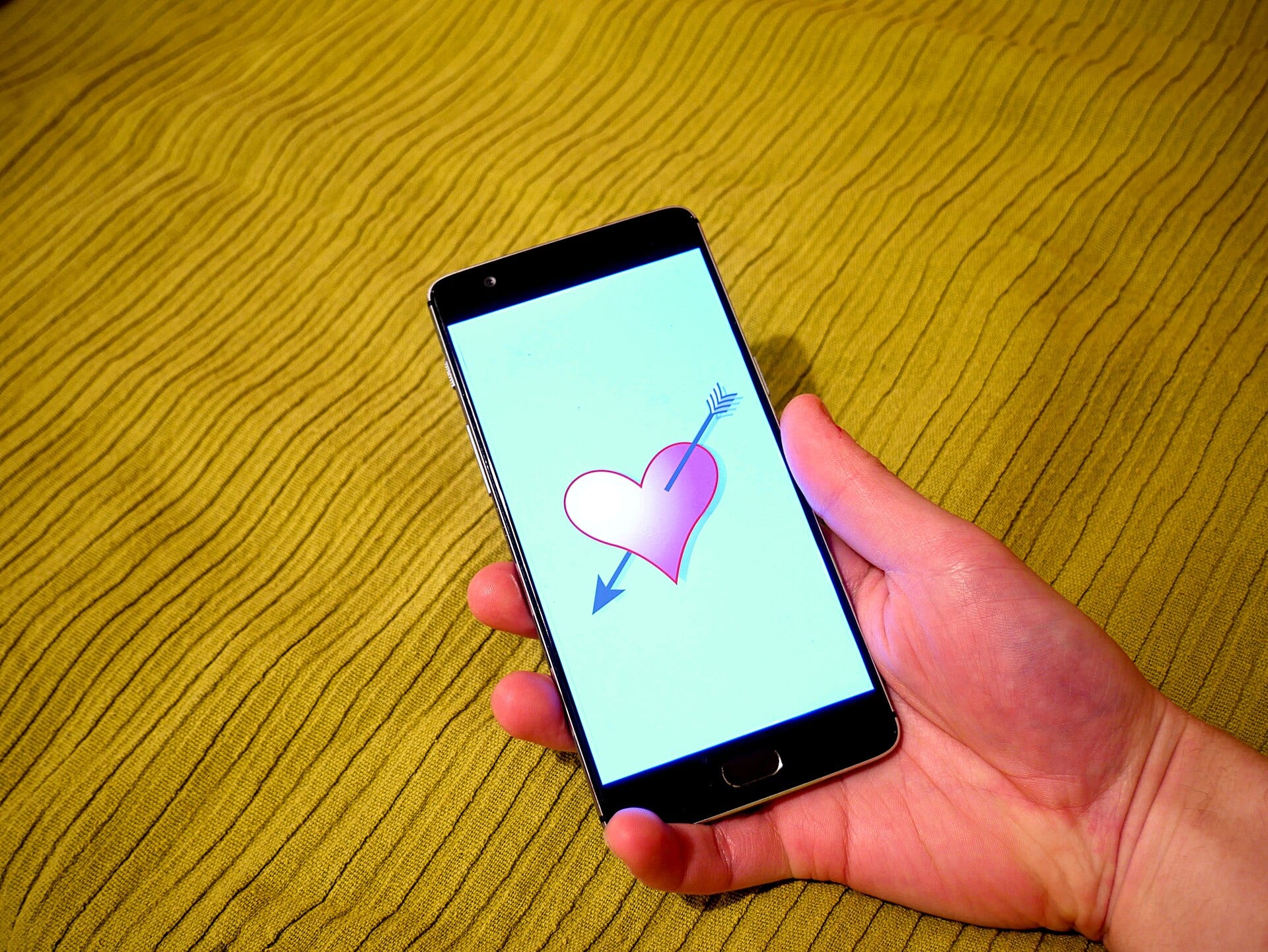 A hand holds a smartphone showing a dating app with a heart pierced by an arrow on its screen