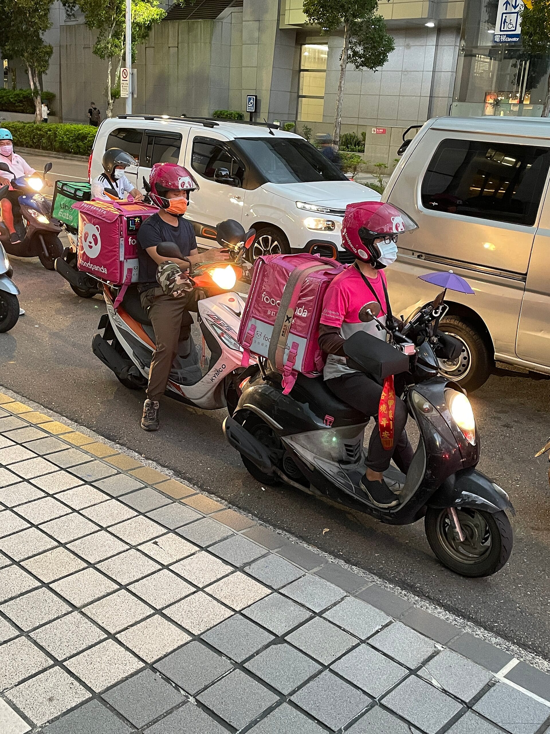 Food delivery couriers on scooters, insulated delivery bags strapped on, wait at a city intersection