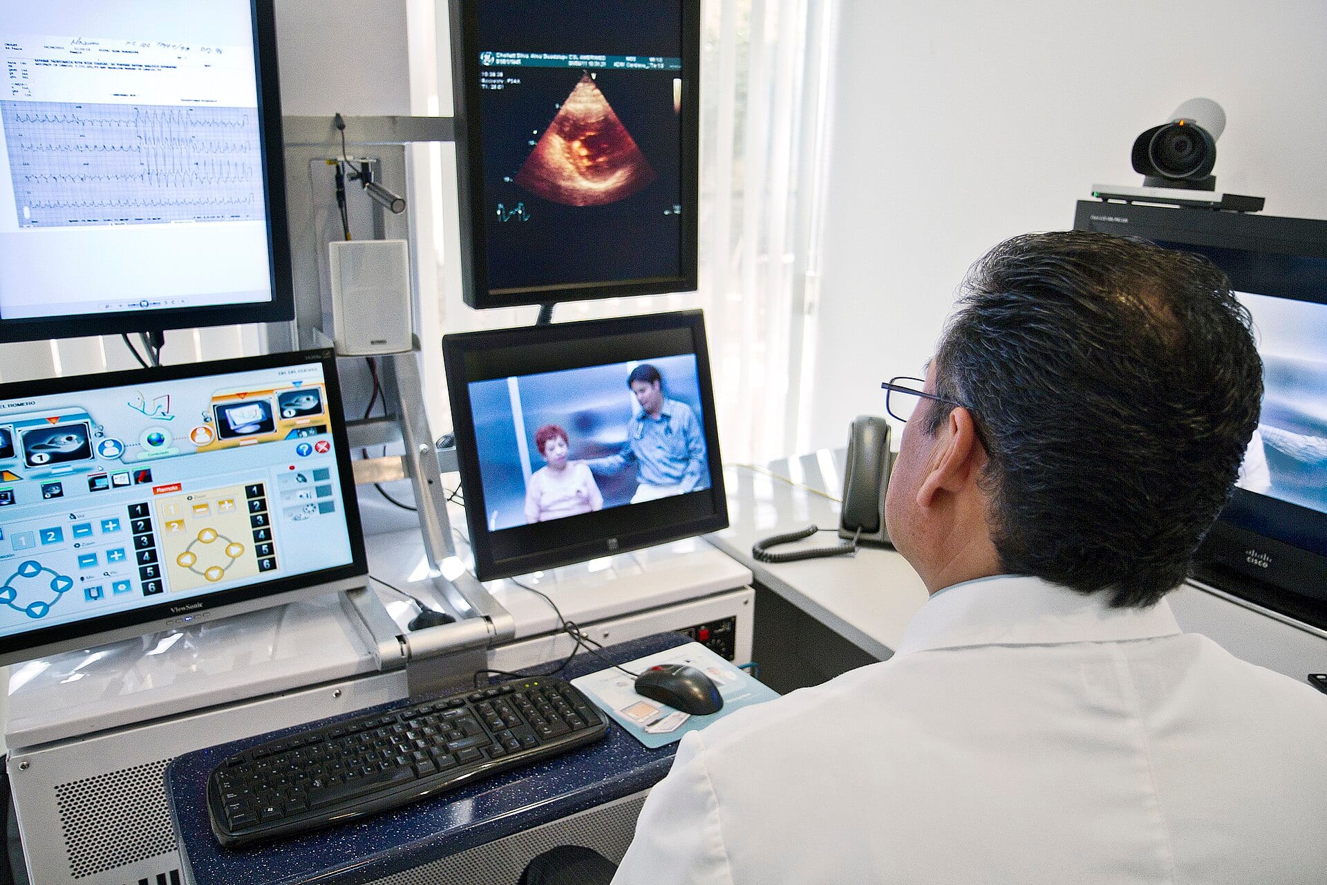 A clinician at a telemedicine workstation consults with a patient by video, with diagnostic readouts on surrounding screens