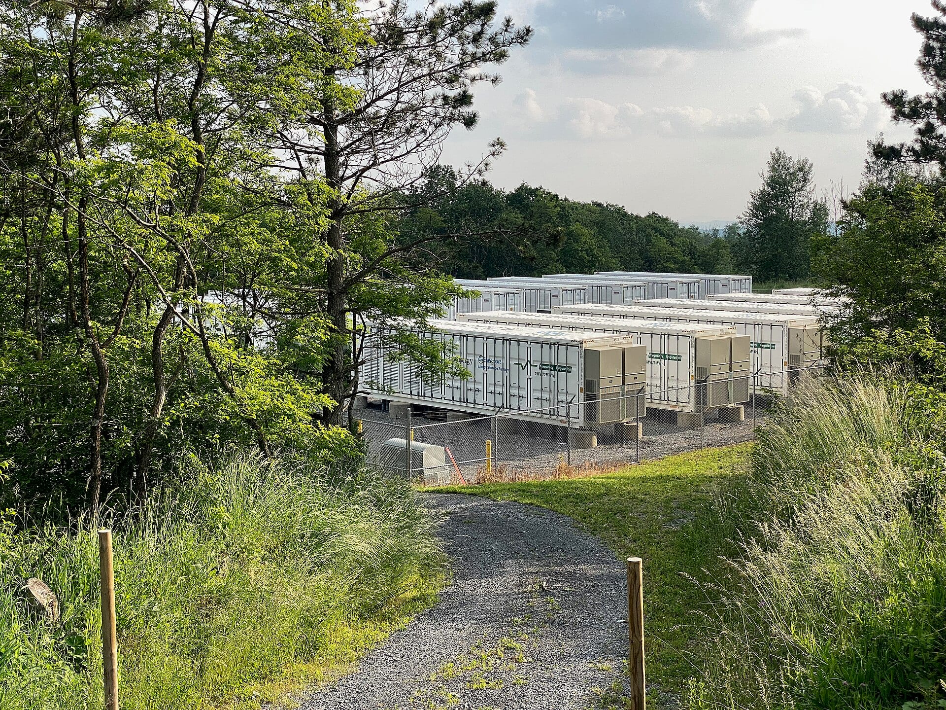 Rows of white battery storage containers behind a chain-link fence at a utility-scale battery energy storage site